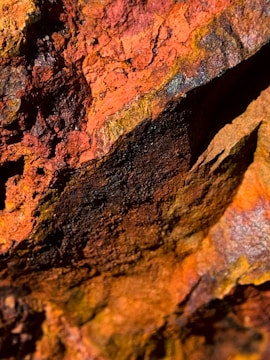 A close-up view of a textured rock surface with shades of orange, red, and black. The rough, uneven texture reveals layered mineral deposits, giving a rugged and weathered appearance.