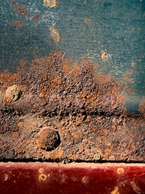 Rusty metal surface with distinct patches of corrosion and textured buildup. Various shades of brown, orange, and dark green intermingle, highlighting the metal's degradation. Rivets and bolt heads can be seen emerging from the corroded areas.