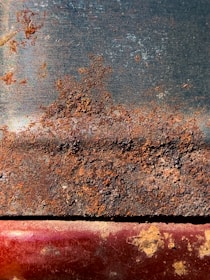 A heavily rusted metal surface displaying a mix of brown and orange corrosion atop a darker, smooth background. The lower part has a hint of red and has patches of rusty textures with some exposed metal areas.
