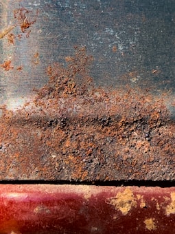 A heavily rusted metal surface displaying a mix of brown and orange corrosion atop a darker, smooth background. The lower part has a hint of red and has patches of rusty textures with some exposed metal areas.