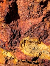A close-up view of a rough, textured surface resembling rust-covered or iron-rich rock, with varying shades of orange, red, and yellow, indicating oxidation or mineral deposits.