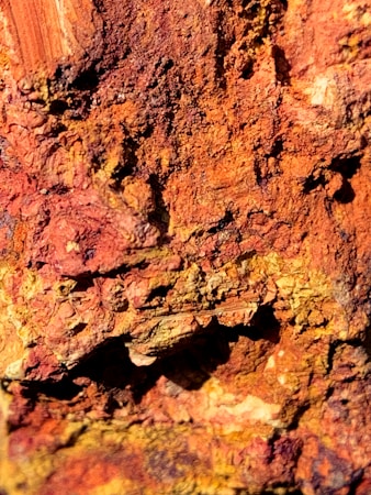 A close-up texture of a rusty, rough surface exhibiting layers of natural erosion. The surface features various shades of reddish-brown, orange, and yellow, indicating oxidation and weathering processes.