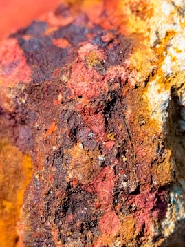 A close-up view of a textured, rocky surface with rich earthy hues, showcasing various shades of red, brown, and yellow. The rugged texture is highlighted by shadows, giving it depth and dimension.