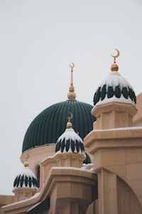 A mosque with ornate domes covered in snow, featuring crescent moon ornaments on top. The structure displays a contrast between the deep green of the domes and the light beige of the building’s walls, creating an elegant and serene atmosphere.