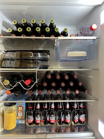 An open refrigerator filled with bottles of beer and other drinks. The top shelf has two stacks of beer bottles lying horizontally, one stack placed next to a transparent storage container. The middle shelf features a wine rack holding various bottles of wine and another stack of beer bottles, vertically aligned. The bottom shelf contains several beer bottles lined up, a jar with an orange liquid, and a blue carton of 100% juice.
