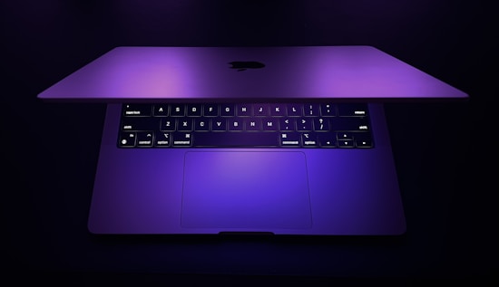 A partially open laptop with an illuminated keyboard. The screen and keys are bathed in a soft purple light, creating a sleek and modern aesthetic.