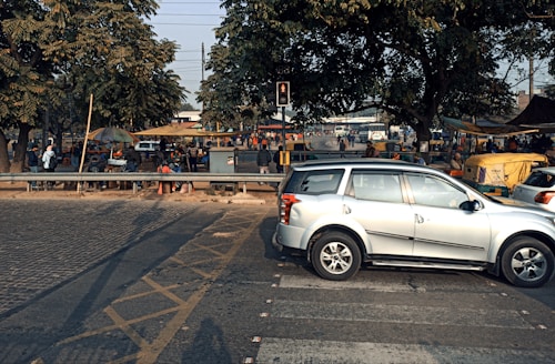 A busy urban street scene featuring a silver SUV stopped at a crosswalk. In the background, several people are gathered around market stalls, some covered with yellow and beige tarps. The area is shaded by large trees, and there are more vehicles and motorcycles visible further back.