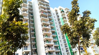 A tall, modern apartment building with multiple floors featuring numerous balconies. The building's facade is decorated with green and white accents. Several trees with lush green leaves are prominently visible in the foreground, enhancing the urban environment.