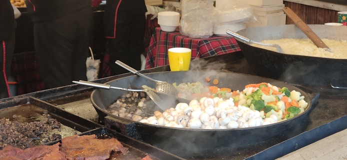 A bustling food stall with large pans of steaming vegetables, including mushrooms, carrots, broccoli, and cauliflower. In the background, there are people in black uniforms and a red and black checkered tablecloth. A yellow paper cup and a bowl are also visible.