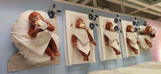 Plush toy monkeys are wrapped in white blankets and mounted on vertical surfaces. They exhibit various expressions and gestures, including drinking from a cup. Blue plush toys and decorative flowers are interspersed among the monkeys.