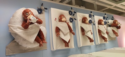 Plush toy monkeys are wrapped in white blankets and mounted on vertical surfaces. They exhibit various expressions and gestures, including drinking from a cup. Blue plush toys and decorative flowers are interspersed among the monkeys.