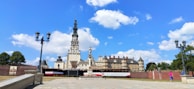A historic religious complex featuring a tall, ornate tower and classical architecture with beige and brown tones. The scene includes a large open square with a few people walking and street lamps. The sky is bright blue with scattered white clouds.