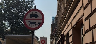 A round red sign with a white background displays black silhouettes of a tractor and a horse-drawn vehicle, indicating a prohibition. The sign is positioned on a street lined with tall buildings and a large tree. Another smaller sign below shows a red circle with a letter 'A' and the word 'Podstrefa.'