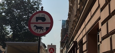A round red sign with a white background displays black silhouettes of a tractor and a horse-drawn vehicle, indicating a prohibition. The sign is positioned on a street lined with tall buildings and a large tree. Another smaller sign below shows a red circle with a letter 'A' and the word 'Podstrefa.'