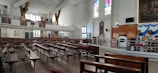 A large, spacious interior with wooden pews arranged in rows. There are colorful stained glass windows above an altar area which is decorated with floral arrangements. The walls and ceiling are painted white, contributing to the brightness of the space. There are also decorative religious artworks and a sound system setup.