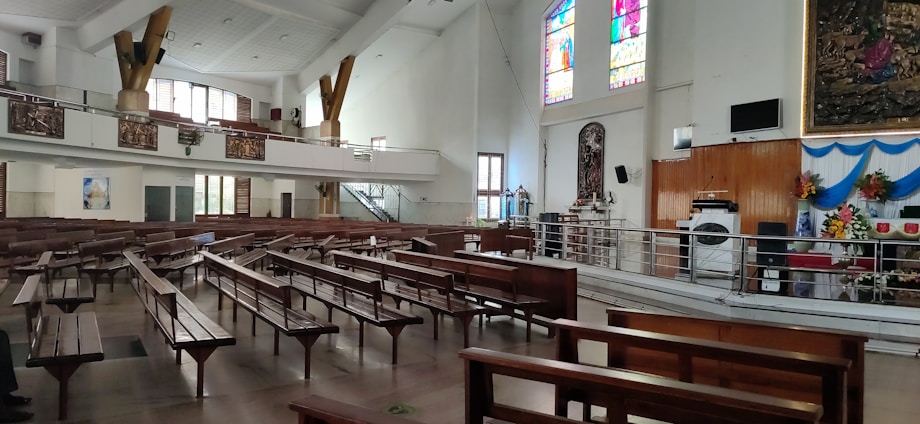 A large, spacious interior with wooden pews arranged in rows. There are colorful stained glass windows above an altar area which is decorated with floral arrangements. The walls and ceiling are painted white, contributing to the brightness of the space. There are also decorative religious artworks and a sound system setup.