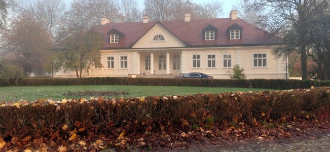 A large, two-story house with a red roof and beige exterior sits in the middle of a well-manicured lawn. There are several trees surrounding the house, with leaves in autumn colors. A black car is parked in front of the house.