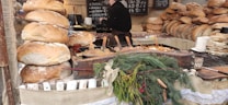A rustic market stall displaying a variety of freshly baked, large round loaves of bread stacked on top of each other. In the foreground, there is a wooden container with different cooked dishes, served with wooden spoons, and garnished with sprigs of pine and berries. Behind the stall, price boards with writing in a foreign language are visible, suggesting a traditional or local market setting.