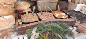 Several large loaves of bread are stacked next to a wooden serving area. There are four pans filled with different cooked dishes, each with a wooden serving spoon. A bundle of evergreen branches and red berries is placed in front of the food. A person is partially visible, standing behind the setup, with stacks of napkins nearby.