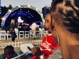 The image captures a close-up view of a person's braided hairstyle and large hoop earring, with a blurred background of people sitting on white chairs. In the distance, a large stage with bright blue and white lights is visible, suggesting a concert or event setting. The focus on the hairstyle and earring adds a personal touch within the bustling atmosphere.