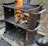 A metal outdoor barbecue grill with handles features a stack of firewood burning brightly. The surrounding area is covered with fallen leaves and the ground is charred. A person's foot is visible near the bottom right corner.