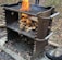 A metal outdoor barbecue grill with handles features a stack of firewood burning brightly. The surrounding area is covered with fallen leaves and the ground is charred. A person's foot is visible near the bottom right corner.