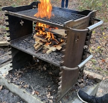 A metal outdoor barbecue grill with handles features a stack of firewood burning brightly. The surrounding area is covered with fallen leaves and the ground is charred. A person's foot is visible near the bottom right corner.