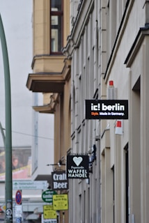 A street view featuring several building facades with multiple shop signs. Notable signs include 'ic! berlin', 'Craft Beer', and 'Waffel Handel'. The architecture is characterized by large windows and a neutral color palette. The street is lined with vertical poles and traffic signs, and there’s a slight bustling urban atmosphere.