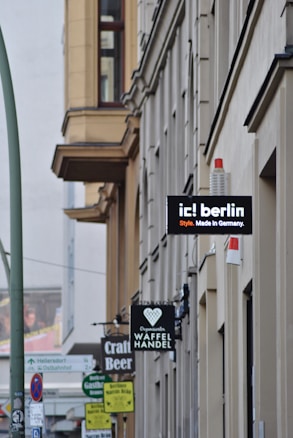A street view featuring several building facades with multiple shop signs. Notable signs include 'ic! berlin', 'Craft Beer', and 'Waffel Handel'. The architecture is characterized by large windows and a neutral color palette. The street is lined with vertical poles and traffic signs, and there&rsquo;s a slight bustling urban atmosphere.