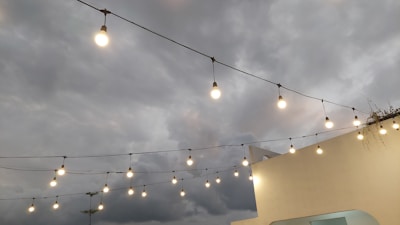 Outdoor string lights are hanging across a cloudy sky, attached to a light-colored wall on the right. The illumination from the bulbs contrasts with the overcast weather, creating a cozy, warm atmosphere despite the gloomy background.