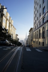 A steep urban street flanked by buildings on both sides, with trolley tracks running down the center. Cars are parked along the street, and in the distance, a distinct pyramid-shaped skyscraper can be seen under a clear blue sky.