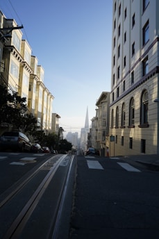 A steep urban street flanked by buildings on both sides, with trolley tracks running down the center. Cars are parked along the street, and in the distance, a distinct pyramid-shaped skyscraper can be seen under a clear blue sky.