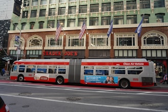 A city street scene featuring a large city bus parked in front of a Trader Joe's store. The bus is a long, articulated vehicle labeled as a 'Clean Air Vehicle' and has advertisements on its side. The building behind the bus is multi-storied with a decorative exterior and several American flags hanging above the entrance.