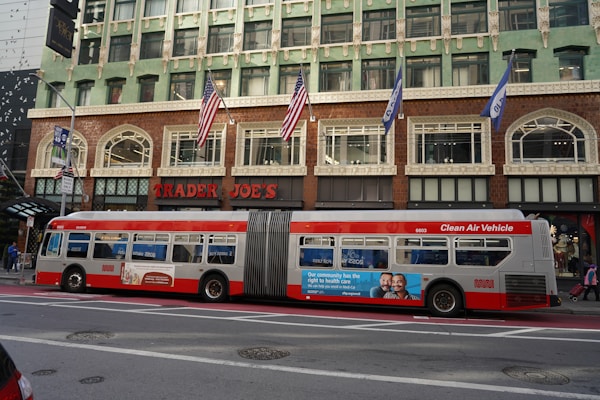 A city street scene featuring a large city bus parked in front of a Trader Joe's store. The bus is a long, articulated vehicle labeled as a 'Clean Air Vehicle' and has advertisements on its side. The building behind the bus is multi-storied with a decorative exterior and several American flags hanging above the entrance.