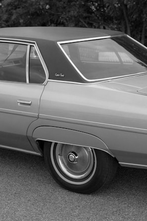 A classic car features prominently, showcasing its distinct design elements such as the sleek roofline and chrome accents. The black and white photo highlights the smooth curves and detailed hubcaps of the vehicle.