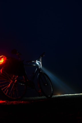 A bicycle stands by the roadside in the dark, illuminated by a bright headlight casting beams into the night. A red tail light is also visible, softly glowing and partially illuminating the rear wheel. The background is a deep dark blue, suggesting it is nighttime or very early morning.