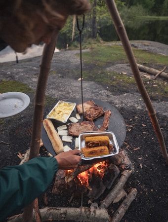 A person is cooking over an open fire with a makeshift grill made of metal supported by wooden poles. The grill holds various foods including corn on the cob, slices of bread, and other grilled items. The setting appears to be outdoors, possibly near a forest or beside a lake, with natural rock and greenery visible in the background.