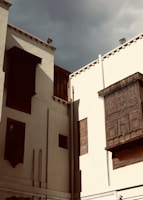 Tall, white-walled buildings with intricate wooden lattice windows stand under a cloudy sky. The architecture showcases Middle Eastern or North African design elements, with decorative mashrabiya screens made of dark wood.