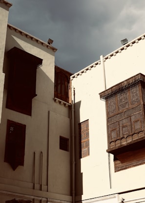 Tall, white-walled buildings with intricate wooden lattice windows stand under a cloudy sky. The architecture showcases Middle Eastern or North African design elements, with decorative mashrabiya screens made of dark wood.