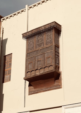 A traditional wooden lattice window protrudes from a beige wall. The intricate geometric patterns and carvings on the window are accentuated by the warm sunlight. The structure showcases heritage architectural elements with its detailed craftsmanship.