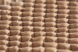 A close-up view of a honeycomb structure made from cardboard, featuring a repeating geometric pattern. The cells are hexagonal and crafted from a light brown paper material, giving a textured appearance with shadows that enhance the depth and structure.
