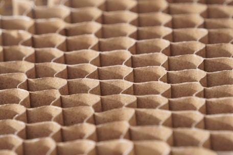 A close-up view of a honeycomb structure made from cardboard, featuring a repeating geometric pattern. The cells are hexagonal and crafted from a light brown paper material, giving a textured appearance with shadows that enhance the depth and structure.