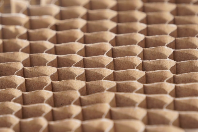 A close-up view of a honeycomb structure made from cardboard, featuring a repeating geometric pattern. The cells are hexagonal and crafted from a light brown paper material, giving a textured appearance with shadows that enhance the depth and structure.