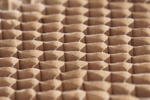 A close-up view of a honeycomb structure made from cardboard, featuring a repeating geometric pattern. The cells are hexagonal and crafted from a light brown paper material, giving a textured appearance with shadows that enhance the depth and structure.