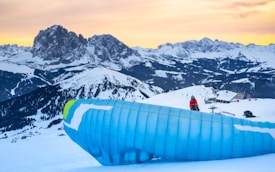 A snowy mountain landscape with a paraglider preparing to take off. The person is dressed in winter gear with a blue helmet, surrounded by snow-covered peaks and a colorful sunset sky. The large blue parachute is prominently displayed in the foreground.