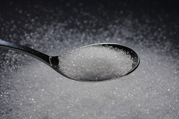 A stainless steel spoon filled with granulated white sugar is placed over a surface covered with additional sugar granules. The fine texture of the sugar crystals catches the light, creating a sparkling effect.