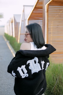 A woman with dark hair and sunglasses is walking along a pathway lined with small, modern wooden structures. She is wearing a white top underneath a black jacket with bold white lettering on the back. Green plants grow alongside the pathway.