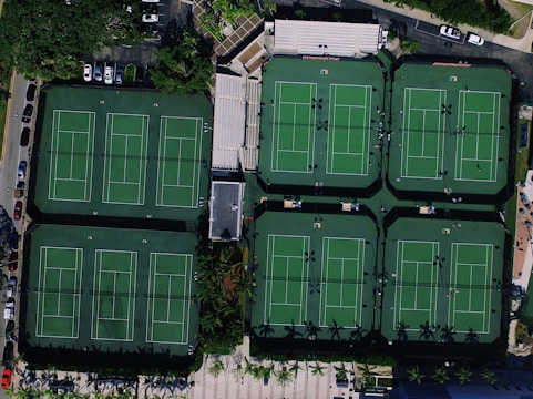 An aerial view of a complex featuring six tennis courts with linear markings, surrounded by parking lots, trees, and seating areas. The courts have a consistent green color, and there are several people visible on some of them, possibly engaging in sports activities.