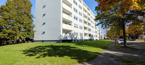 A multi-story residential building with a white facade is situated next to a lush green lawn bordered by a row of trees displaying autumn foliage. A street runs parallel to the building with several parked cars. The sky is clear, enhancing the brightness of the scene.