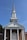 A traditional church building with a tall steeple against a clear blue sky, featuring a brick facade and white columns. The words 'CHURCH OF CHRIST' are prominently displayed above the entrance.